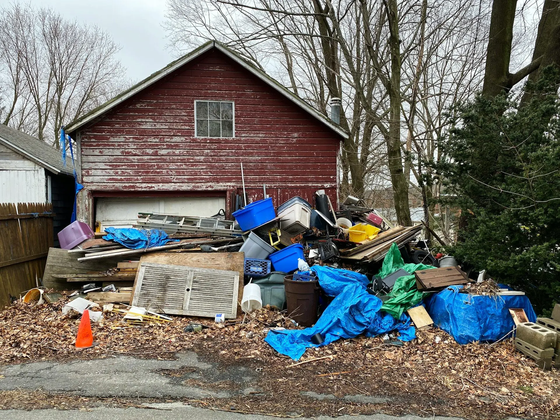 Large pile of household debris, plastic tubs, blue tarps, wooden waste stacked outside red garage shed