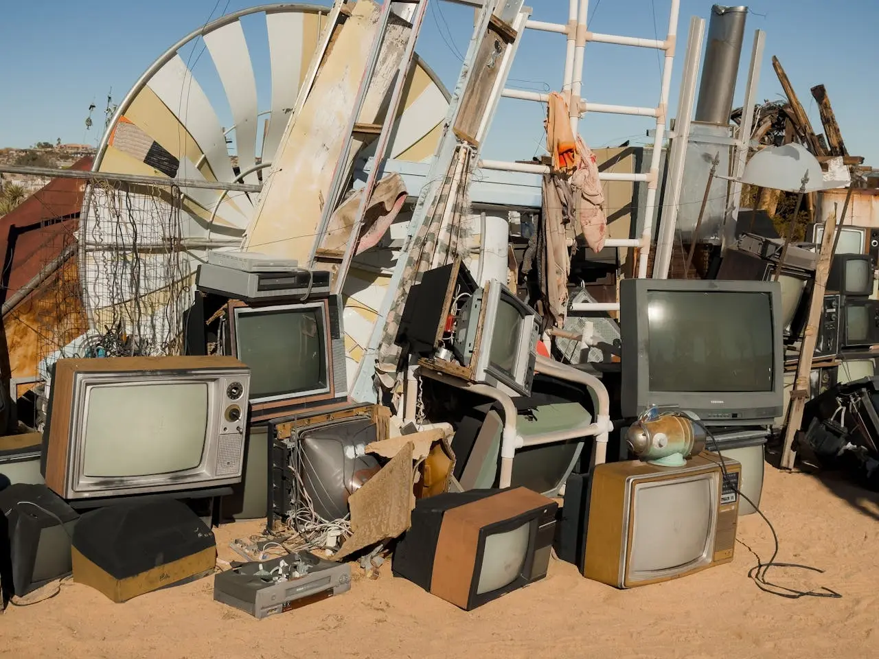 Large stack of old television sets, electronic waste, ladders, scrap metal cluttering outdoors on dirt ground