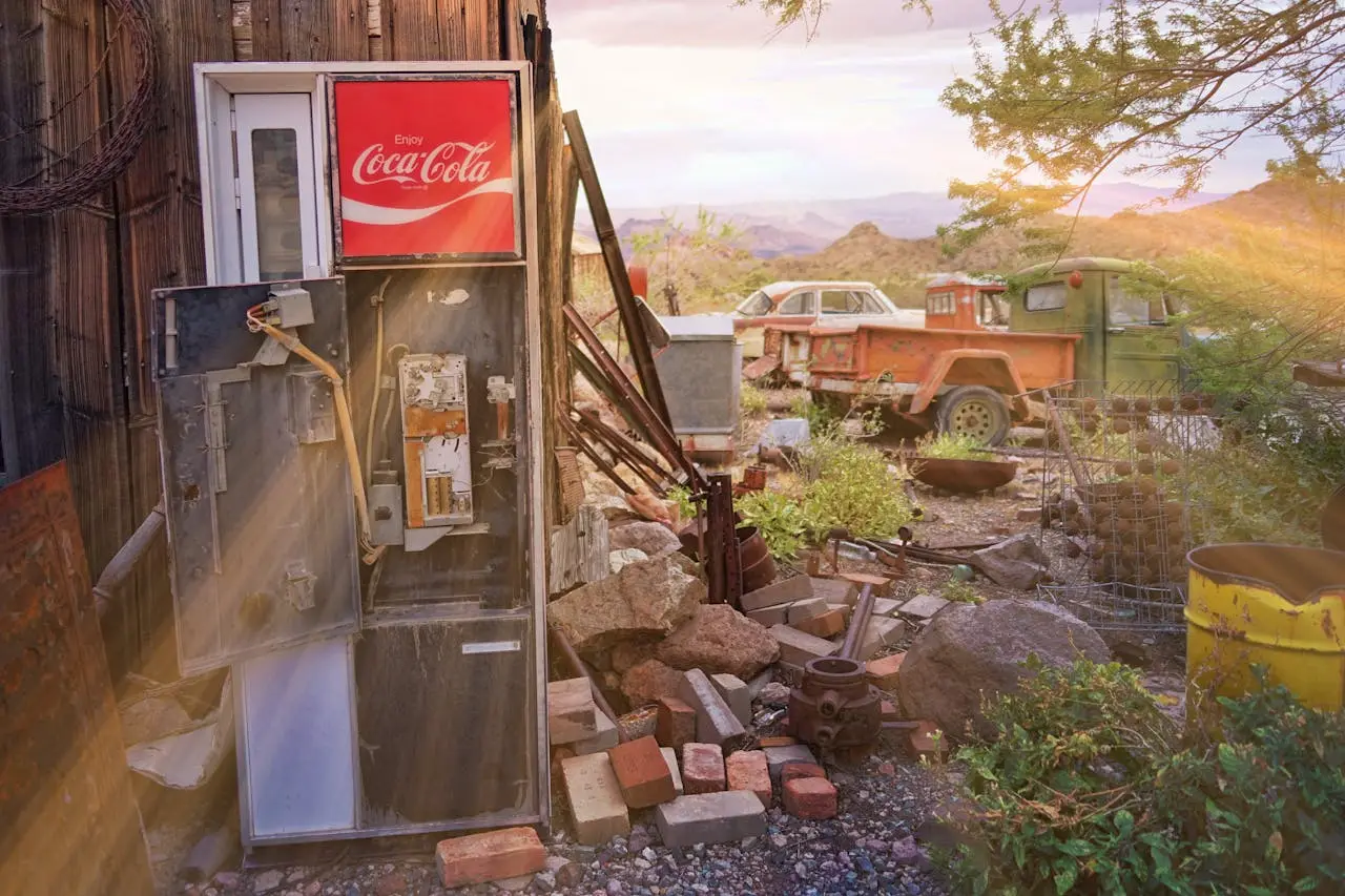 Old vintage Coca-Cola vending machine, rusted truck, abandoned scrap metal items in outdoor junkyard